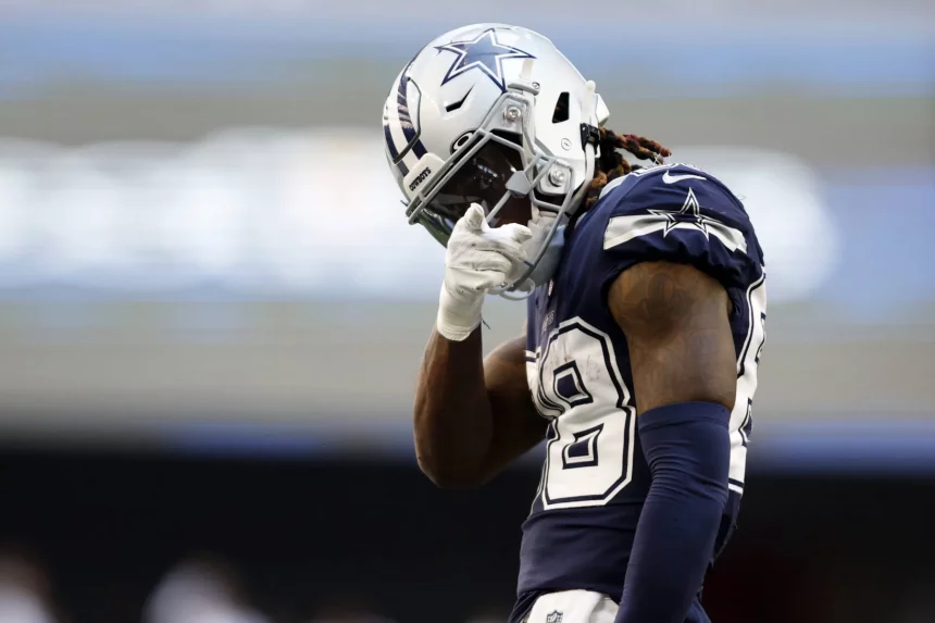 Ceedee Lamb im Trikot der Dallas Cowboys steht auf dem Spielfeld und vollführt die klassische "Nasenwischer" Geste. Er trägt ein marineblaues Trikot mit weißen Akzenten, die Nummer 88, einen silbernen Helm und weiße Handschuhe und rückt konzentriert seinen Helm zurecht – ein Zeichen seiner Hingabe an die NFL-Regeln. Diese Beschreibung wurde mit der FootballR KI automatisch generiert.