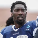 Ein Footballspieler, der an DeMarcus Lawrence erinnert, trägt ein blau-weißes Trikot mit der Aufschrift „AT&T“ auf der Vorderseite und blickt zur Seite. Er hat kurzes, lockiges Haar und einen Bart vor einem unscharfen Hintergrund, der auf undeutliche Gestalten hindeutet, wahrscheinlich Teamkollegen oder andere Personen. Diese Beschreibung wurde mit der FootballR KI automatisch generiert.