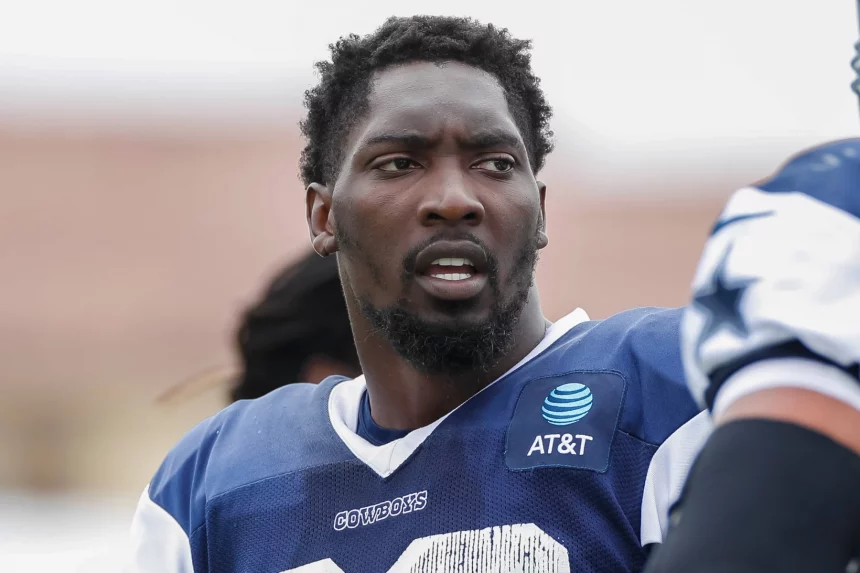 Ein Footballspieler, der an DeMarcus Lawrence erinnert, trägt ein blau-weißes Trikot mit der Aufschrift „AT&T“ auf der Vorderseite und blickt zur Seite. Er hat kurzes, lockiges Haar und einen Bart vor einem unscharfen Hintergrund, der auf undeutliche Gestalten hindeutet, wahrscheinlich Teamkollegen oder andere Personen. Diese Beschreibung wurde mit der FootballR KI automatisch generiert.