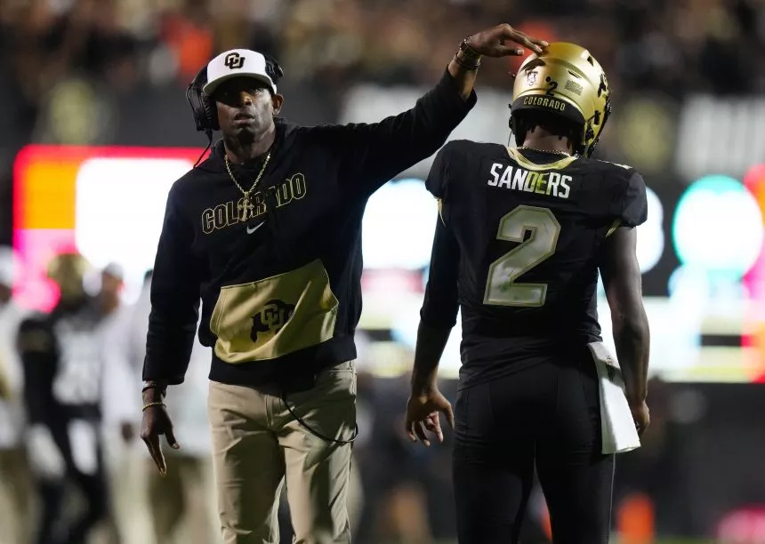 Deion Sanders in schwarzem Colorado-Hoodie und weißer Kappe deutet auf Shedeur Sanders, der eine schwarze Uniform mit der Nummer 2 trägt. Sein Helm glänzt golden mit der Aufschrift „Colorado“. Sie stehen auf dem Spielfeld, umgeben von verschwommenen Zuschauern und hellen Stadionlichtern. Diese Beschreibung wurde mit der FootballR KI automatisch generiert.