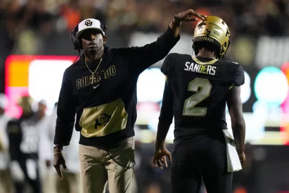 Deion Sanders in schwarzem Colorado-Hoodie und weißer Kappe deutet auf Shedeur Sanders, der eine schwarze Uniform mit der Nummer 2 trägt. Sein Helm glänzt golden mit der Aufschrift „Colorado“. Sie stehen auf dem Spielfeld, umgeben von verschwommenen Zuschauern und hellen Stadionlichtern. Diese Beschreibung wurde mit der FootballR KI automatisch generiert.