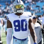 DeMarcus Lawrence, Footballspieler der Dallas Cowboys, steht auf dem Spielfeld. Er trägt ein weißes Trikot mit der Nummer 90, weiße Hosen und eine verspiegelte Sonnenbrille. Im Hintergrund sind Teamkollegen und Trainer zu sehen, dazu ein Stadion voller Zuschauer. Diese Beschreibung wurde mit der FootballR KI automatisch generiert.