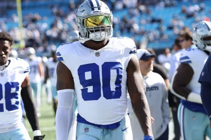 DeMarcus Lawrence, Footballspieler der Dallas Cowboys, steht auf dem Spielfeld. Er trägt ein weißes Trikot mit der Nummer 90, weiße Hosen und eine verspiegelte Sonnenbrille. Im Hintergrund sind Teamkollegen und Trainer zu sehen, dazu ein Stadion voller Zuschauer. Diese Beschreibung wurde mit der FootballR KI automatisch generiert.