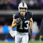 Hunter Renfrow in einem dunklen Trikot mit der Nummer 13, das an Hunter Renfrows Stil erinnert, sprintet mit einem Football in der Hand über das Feld. Er trägt einen silbernen Helm und eine schwarze Hose. Der unscharfe Hintergrund zeigt Zuschauer am Spielfeldrand inmitten der lebhaften Atmosphäre des Stadions. Diese Beschreibung wurde mit der FootballR KI automatisch generiert.
