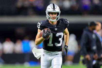 Hunter Renfrow in einem dunklen Trikot mit der Nummer 13, das an Hunter Renfrows Stil erinnert, sprintet mit einem Football in der Hand über das Feld. Er trägt einen silbernen Helm und eine schwarze Hose. Der unscharfe Hintergrund zeigt Zuschauer am Spielfeldrand inmitten der lebhaften Atmosphäre des Stadions. Diese Beschreibung wurde mit der FootballR KI automatisch generiert.