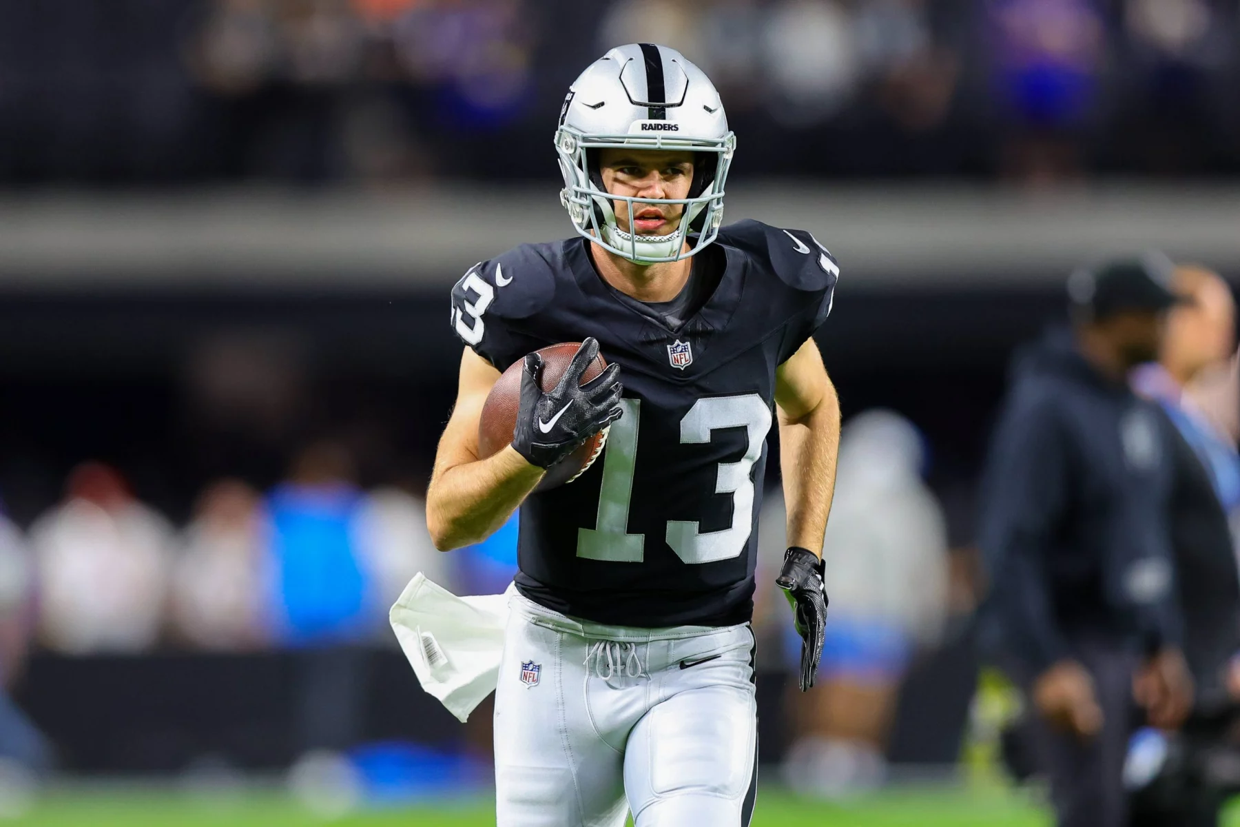 Hunter Renfrow in einem dunklen Trikot mit der Nummer 13, das an Hunter Renfrows Stil erinnert, sprintet mit einem Football in der Hand über das Feld. Er trägt einen silbernen Helm und eine schwarze Hose. Der unscharfe Hintergrund zeigt Zuschauer am Spielfeldrand inmitten der lebhaften Atmosphäre des Stadions. Diese Beschreibung wurde mit der FootballR KI automatisch generiert.