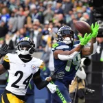 Ein Footballspieler in blau-grüner Uniform, die an DK Metcalf Stil erinnert, fängt einen Ball in der Luft und streckt sich nach vorne, während ein Spieler in Schwarz-Gelb – möglicherweise von den Steelers – ihn dicht verteidigt. Die Menge beobachtet die spannende Action. Diese Beschreibung wurde mit der FootballR KI automatisch generiert.