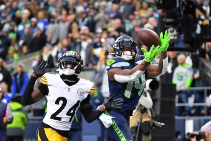 Ein Footballspieler in blau-grüner Uniform, die an DK Metcalf Stil erinnert, fängt einen Ball in der Luft und streckt sich nach vorne, während ein Spieler in Schwarz-Gelb – möglicherweise von den Steelers – ihn dicht verteidigt. Die Menge beobachtet die spannende Action. Diese Beschreibung wurde mit der FootballR KI automatisch generiert.