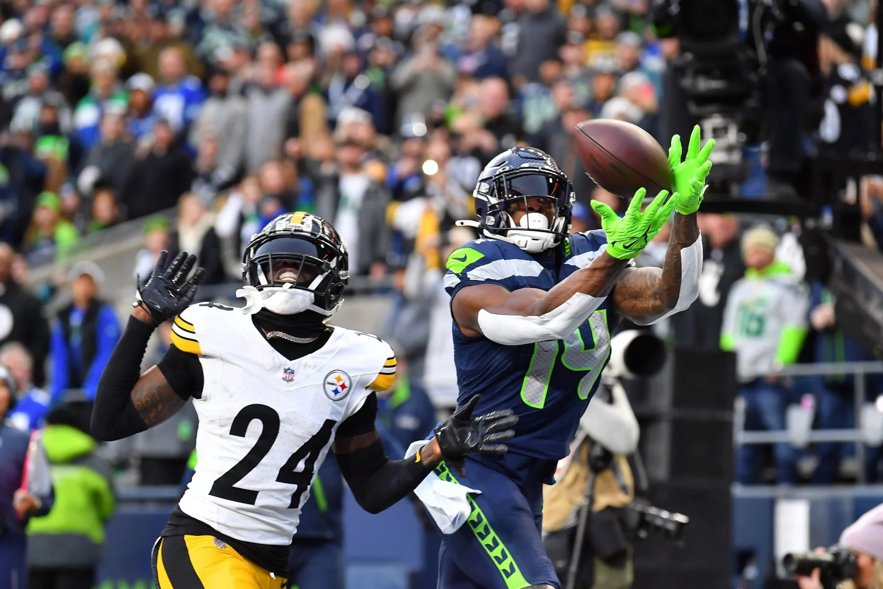 Ein Footballspieler in blau-grüner Uniform, die an DK Metcalf Stil erinnert, fängt einen Ball in der Luft und streckt sich nach vorne, während ein Spieler in Schwarz-Gelb – möglicherweise von den Steelers – ihn dicht verteidigt. Die Menge beobachtet die spannende Action. Diese Beschreibung wurde mit der FootballR KI automatisch generiert.