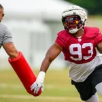 Jonathan Allen im roten Trikot mit der Nummer 93, der Jonathan Allen ähnelt, trainiert auf einem Feld. Er stemmt sich gegen eine rote Blockiermatte, die von einem Trainer in grauer Kleidung gehalten wird. Die Szene spielt sich auf einer Wiese mit verschwommenen Strukturen im Hintergrund ab. Diese Beschreibung wurde mit der FootballR KI automatisch generiert.