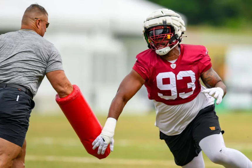 Jonathan Allen im roten Trikot mit der Nummer 93, der Jonathan Allen ähnelt, trainiert auf einem Feld. Er stemmt sich gegen eine rote Blockiermatte, die von einem Trainer in grauer Kleidung gehalten wird. Die Szene spielt sich auf einer Wiese mit verschwommenen Strukturen im Hintergrund ab. Diese Beschreibung wurde mit der FootballR KI automatisch generiert.