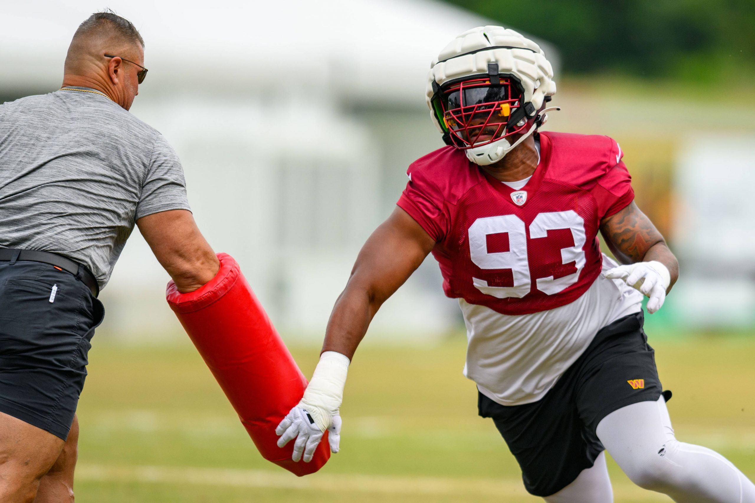 Jonathan Allen im roten Trikot mit der Nummer 93, der Jonathan Allen ähnelt, trainiert auf einem Feld. Er stemmt sich gegen eine rote Blockiermatte, die von einem Trainer in grauer Kleidung gehalten wird. Die Szene spielt sich auf einer Wiese mit verschwommenen Strukturen im Hintergrund ab. Diese Beschreibung wurde mit der FootballR KI automatisch generiert.
