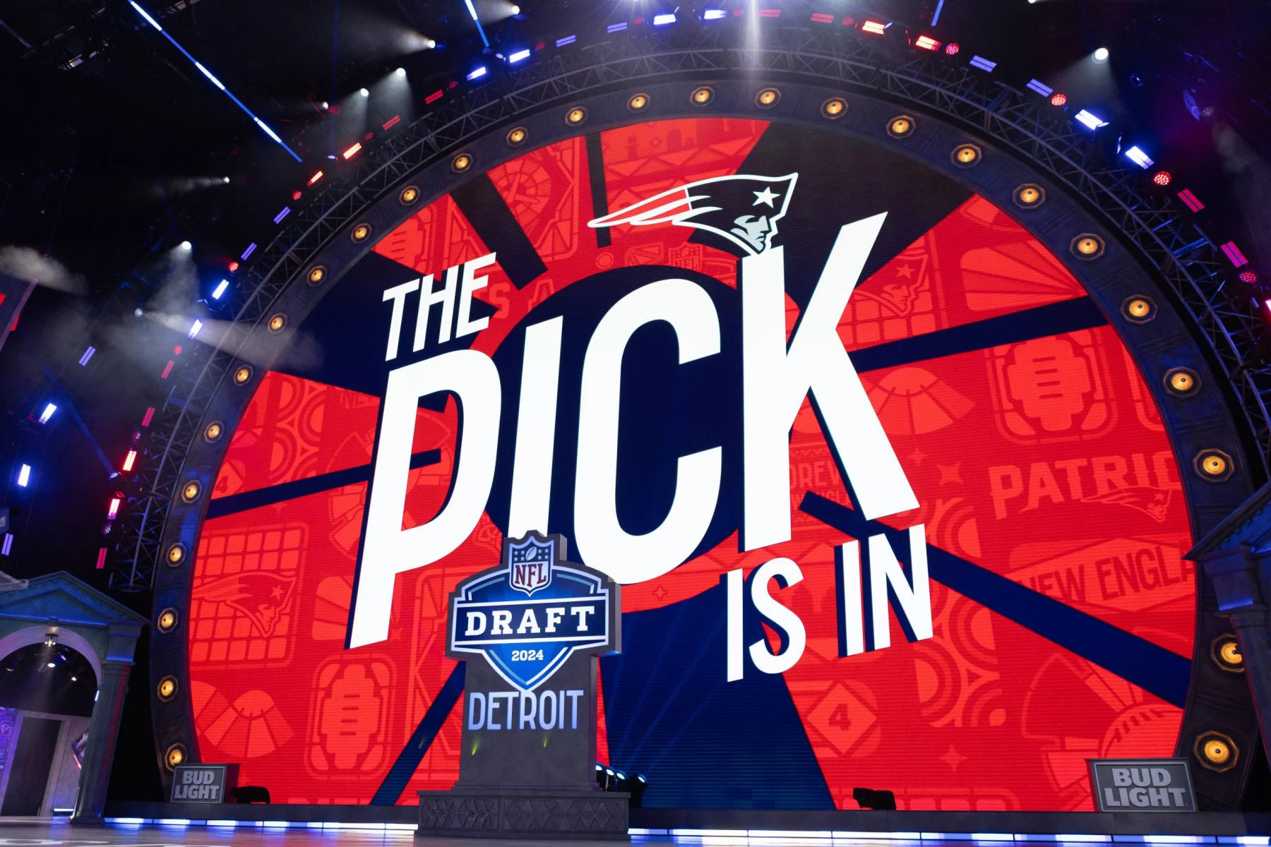 Compensatory Picks 2026 -Auf einer großen Bühne mit einem runden LED-Bildschirm wird neben dem Logo der New England Patriots „The Pick is In“ gezeigt. Football-Symbole schmücken den Hintergrund, während Hinweise auf den NFL Draft 2024 in Detroit und das Bud Light-Branding diesen spannenden Moment abrunden. Diese Beschreibung wurde mit der FootballR KI automatisch generiert.