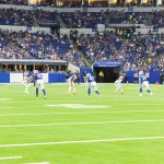 Der spannende Kickoff des Footballspiels findet in einem lebendigen Stadion mit üppigem Grün statt. Spieler in blauen Trikots stehen ihren weißgekleideten Gegnern an der Line of Scrimmage unter hellem Stadionlicht gegenüber. Zuschauer in blauen Trikots füllen die Tribünen, während „Caesars Rewards“ sie mit Sicherheit und Spannung begrüßt. Diese Beschreibung wurde mit der FootballR KI automatisch generiert.