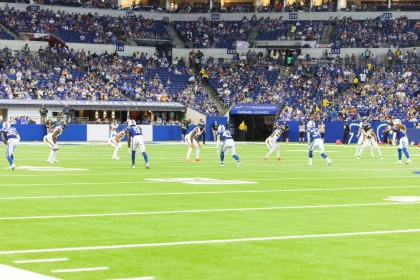 Der spannende Kickoff des Footballspiels findet in einem lebendigen Stadion mit üppigem Grün statt. Spieler in blauen Trikots stehen ihren weißgekleideten Gegnern an der Line of Scrimmage unter hellem Stadionlicht gegenüber. Zuschauer in blauen Trikots füllen die Tribünen, während „Caesars Rewards“ sie mit Sicherheit und Spannung begrüßt. Diese Beschreibung wurde mit der FootballR KI automatisch generiert.