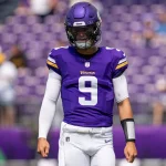 Ein Footballspieler, der J.J. McCarthy ähnelt, trägt ein lila Trikot mit der Nummer 9 auf dem Spielfeld. Zu seiner Ausrüstung gehören ein lila Helm mit dem Teamlogo, weiße Hosen und lange weiße Ärmel. Im Stadionhintergrund sitzen verschwommene Zuschauer auf passenden lila Sitzen. Diese Beschreibung wurde mit der FootballR KI automatisch generiert.