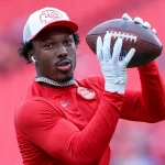 Wide Receiver Mecole Hardman in einem roten Langarmshirt und einer weiß-roten Kappe bereitet sich darauf vor, einen Football zu fangen. Mit weißen Handschuhen, einer Halskette und Ohrstöpseln steht er vor einer verschwommenen roten Stadionkulisse. Diese Beschreibung wurde mit der FootballR KI automatisch generiert.