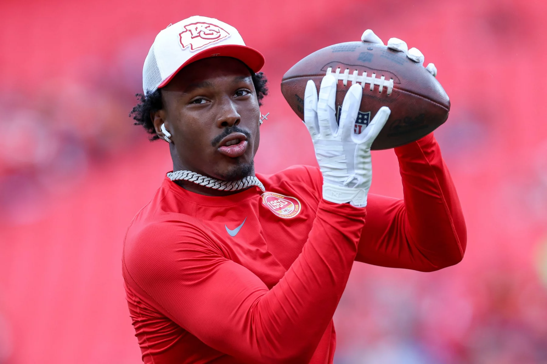 Wide Receiver Mecole Hardman in einem roten Langarmshirt und einer weiß-roten Kappe bereitet sich darauf vor, einen Football zu fangen. Mit weißen Handschuhen, einer Halskette und Ohrstöpseln steht er vor einer verschwommenen roten Stadionkulisse. Diese Beschreibung wurde mit der FootballR KI automatisch generiert.