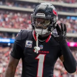 Stefon Diggs in der schwarzen Houston-Texans-Uniform, die an Stefon Diggs' unverwechselbaren Stil erinnert, gestikuliert mit der rechten Hand am Ohr. Er steht auf dem Spielfeld, die Nummer 1 und „TEXANS“ prangen auf seiner Brust, im Hintergrund prangt stolz ein Kapitänsabzeichen inmitten einer verschwommenen Menschenmenge. Diese Beschreibung wurde mit der FootballR KI automatisch generiert.