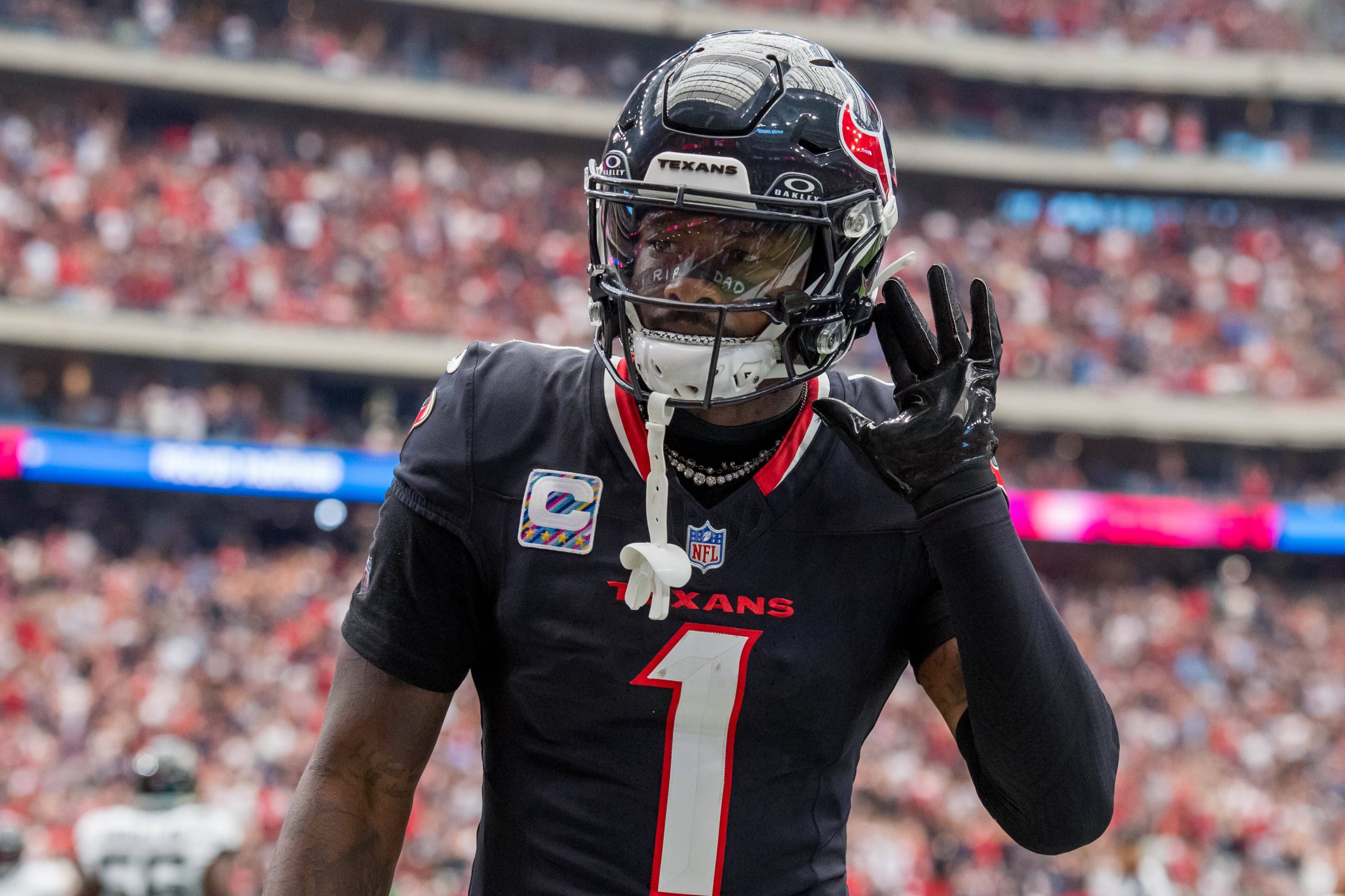 Stefon Diggs in der schwarzen Houston-Texans-Uniform, die an Stefon Diggs' unverwechselbaren Stil erinnert, gestikuliert mit der rechten Hand am Ohr. Er steht auf dem Spielfeld, die Nummer 1 und „TEXANS“ prangen auf seiner Brust, im Hintergrund prangt stolz ein Kapitänsabzeichen inmitten einer verschwommenen Menschenmenge. Diese Beschreibung wurde mit der FootballR KI automatisch generiert.