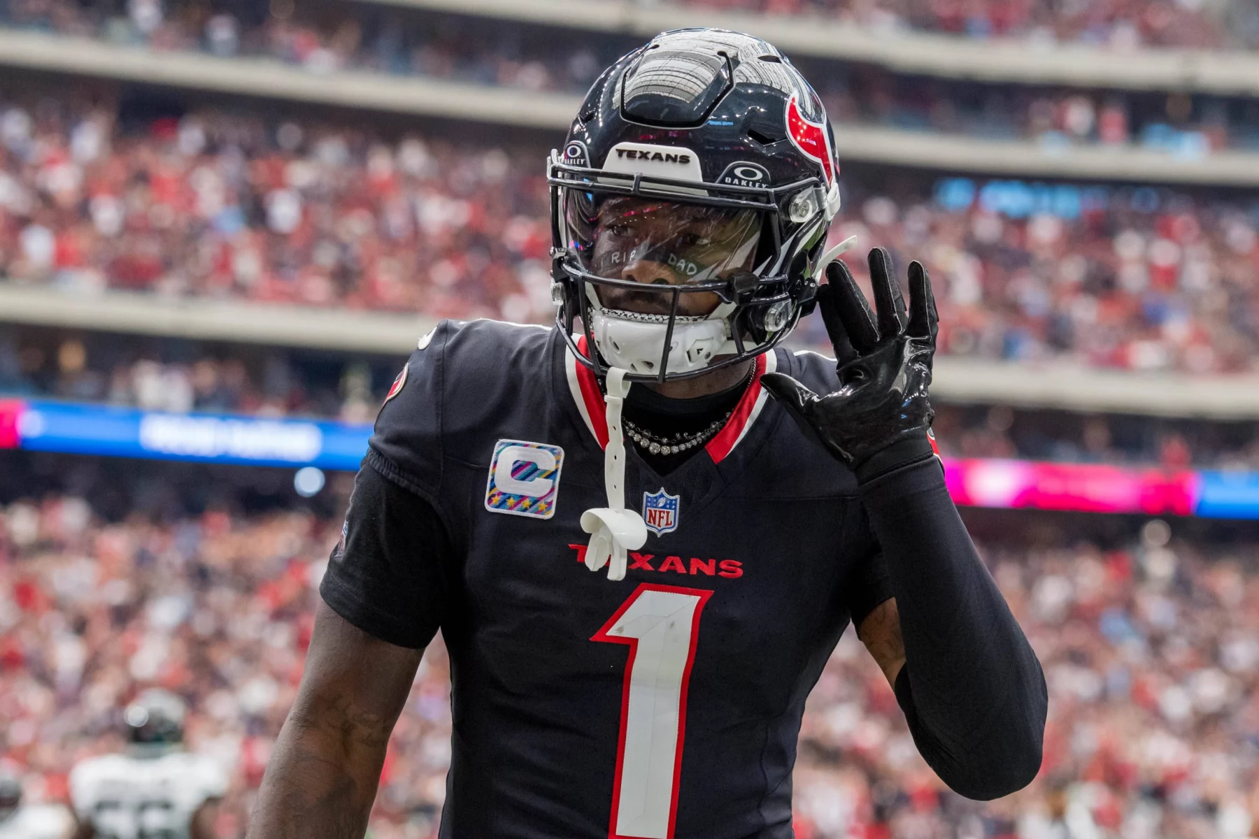 Stefon Diggs in der schwarzen Houston-Texans-Uniform, die an Stefon Diggs' unverwechselbaren Stil erinnert, gestikuliert mit der rechten Hand am Ohr. Er steht auf dem Spielfeld, die Nummer 1 und „TEXANS“ prangen auf seiner Brust, im Hintergrund prangt stolz ein Kapitänsabzeichen inmitten einer verschwommenen Menschenmenge. Diese Beschreibung wurde mit der FootballR KI automatisch generiert.