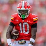 Footballspieler Tai Felton trägt ein rotes Trikot mit der Nummer 10 und dazu passenden Helm und hält einen Football in der Hand. Seine Uniform weist schwarze, gelbe und weiße Akzente auf. Er steht auf einem unscharfen Spielfeld mit seinen Teamkollegen im Hintergrund, was eine aktive Spielumgebung darstellt. Diese Beschreibung wurde mit der FootballR KI automatisch generiert.