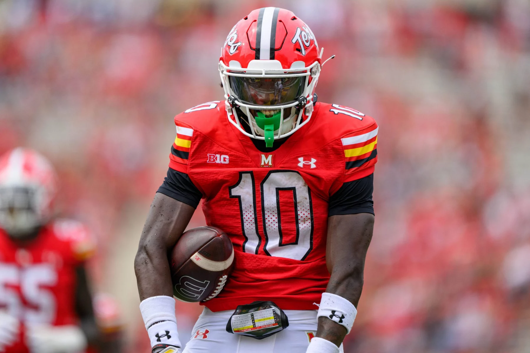 Footballspieler Tai Felton trägt ein rotes Trikot mit der Nummer 10 und dazu passenden Helm und hält einen Football in der Hand. Seine Uniform weist schwarze, gelbe und weiße Akzente auf. Er steht auf einem unscharfen Spielfeld mit seinen Teamkollegen im Hintergrund, was eine aktive Spielumgebung darstellt. Diese Beschreibung wurde mit der FootballR KI automatisch generiert.