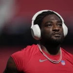 Brandon Aiyuk in einem roten ärmellosen San Francisco 49ers Trikot und zwei Ketten steht auf dem Spielfeld. Er trägt große weiße Kopfhörer. Der Hintergrund ist unscharf und vermittelt Stadionatmosphäre. Der Gesichtsausdruck des Spielers wirkt konzentriert und nachdenklich. Diese Beschreibung wurde mit der FootballR KI automatisch generiert.
