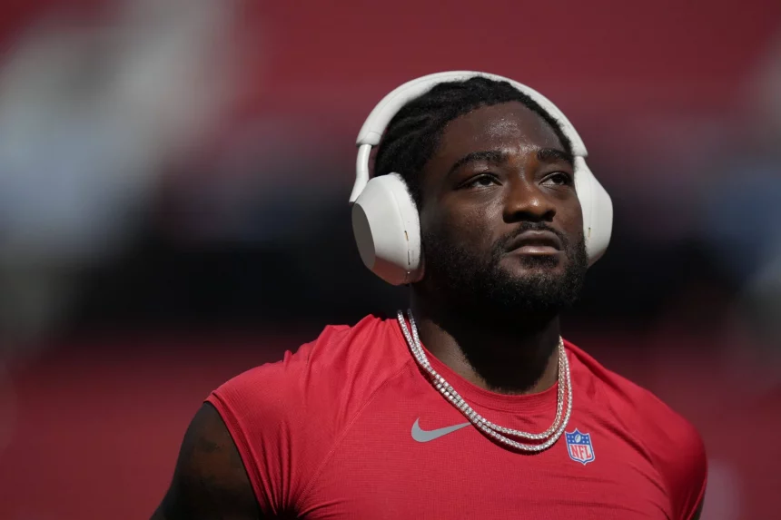 Brandon Aiyuk in einem roten ärmellosen San Francisco 49ers Trikot und zwei Ketten steht auf dem Spielfeld. Er trägt große weiße Kopfhörer. Der Hintergrund ist unscharf und vermittelt Stadionatmosphäre. Der Gesichtsausdruck des Spielers wirkt konzentriert und nachdenklich. Diese Beschreibung wurde mit der FootballR KI automatisch generiert.