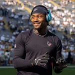 Stefon Diggs steht in einem schwarzen Langarmshirt mit Logo und Handschuhen auf dem Feld. Mit blauen Kopfhörern und Stirnband lächelt er nach links. Die verschwommene Menge füllt die gelben Tribünen des Stadions und sorgt für eine elektrisierende Atmosphäre. Diese Beschreibung wurde mit der FootballR KI automatisch generiert.