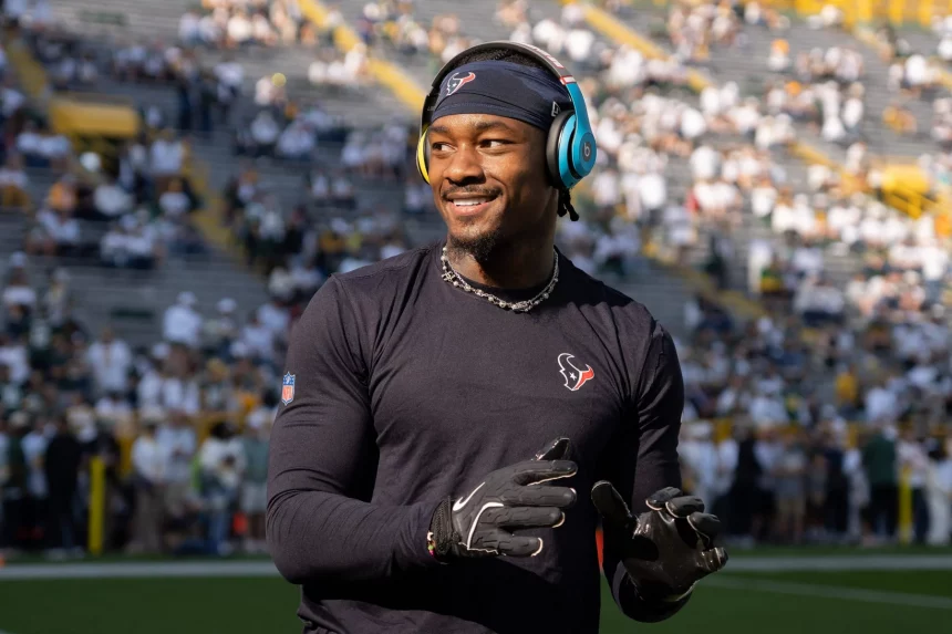 Stefon Diggs steht in einem schwarzen Langarmshirt mit Logo und Handschuhen auf dem Feld. Mit blauen Kopfhörern und Stirnband lächelt er nach links. Die verschwommene Menge füllt die gelben Tribünen des Stadions und sorgt für eine elektrisierende Atmosphäre. Diese Beschreibung wurde mit der FootballR KI automatisch generiert.