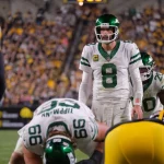 Quarterback Aaron Rodgers in weißem Trikot mit der Nummer 8 steht hinter seinen Teamkollegen und ahmt die souveräne Haltung von Aaron Rodgers während eines Spiels nach. Gegenüber stehen ihm gegnerische Spieler in schwarz-gelben Trikots gegenüber, während im Hintergrund das Stadion von Zuschauern wimmelt. Diese Beschreibung wurde mit der FootballR KI automatisch generiert.