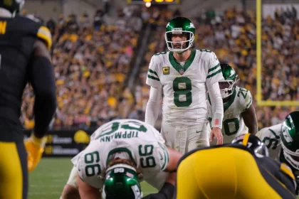 Quarterback Aaron Rodgers in weißem Trikot mit der Nummer 8 steht hinter seinen Teamkollegen und ahmt die souveräne Haltung von Aaron Rodgers während eines Spiels nach. Gegenüber stehen ihm gegnerische Spieler in schwarz-gelben Trikots gegenüber, während im Hintergrund das Stadion von Zuschauern wimmelt. Diese Beschreibung wurde mit der FootballR KI automatisch generiert.