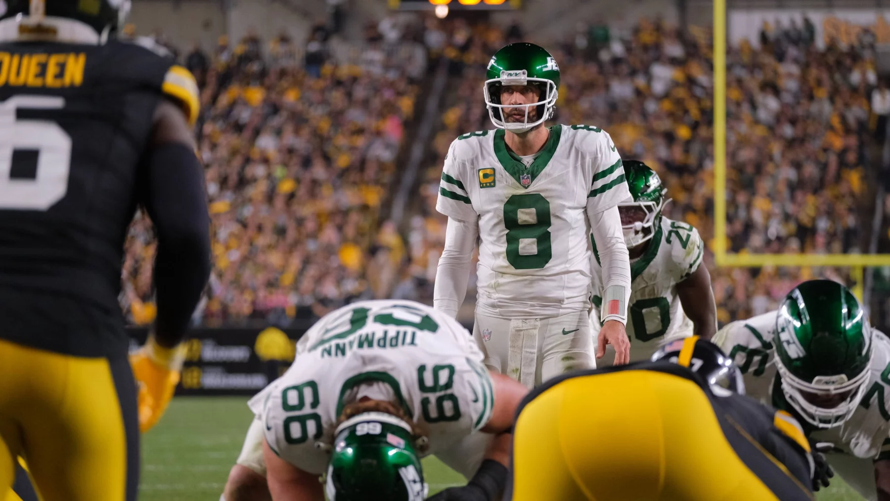 Quarterback Aaron Rodgers in weißem Trikot mit der Nummer 8 steht hinter seinen Teamkollegen und ahmt die souveräne Haltung von Aaron Rodgers während eines Spiels nach. Gegenüber stehen ihm gegnerische Spieler in schwarz-gelben Trikots gegenüber, während im Hintergrund das Stadion von Zuschauern wimmelt. Diese Beschreibung wurde mit der FootballR KI automatisch generiert.