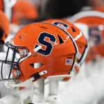 Colorado und Syracuse - Nahaufnahme von leuchtend orangefarbenen Footballhelmen mit einem großen marineblauen „S“-Logo, das an das ikonische Design von Syracuse erinnert, auf einer Bank. Die Helme haben schwarze Gesichtsmasken und einen Aufkleber mit der amerikanischen Flagge auf der Rückseite. Spieler in orange-weißen Trikots sind im Hintergrund leicht unscharf zu sehen. Diese Beschreibung wurde mit der FootballR KI automatisch generiert.