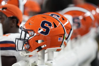 Colorado und Syracuse - Nahaufnahme von leuchtend orangefarbenen Footballhelmen mit einem großen marineblauen „S“-Logo, das an das ikonische Design von Syracuse erinnert, auf einer Bank. Die Helme haben schwarze Gesichtsmasken und einen Aufkleber mit der amerikanischen Flagge auf der Rückseite. Spieler in orange-weißen Trikots sind im Hintergrund leicht unscharf zu sehen. Diese Beschreibung wurde mit der FootballR KI automatisch generiert.