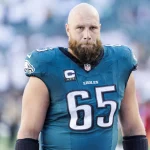 Lane Johnson in einem blaugrünen Trikot der Philadelphia Eagles mit der Nummer 65, vermutlich Lane Johnson, steht auf dem Spielfeld. Er hat einen dichten Bart und eine Glatze. Auf der Uniform des Spielers ist ein Kapitänsabzeichen zu sehen. Im Hintergrund sind eine verschwommene Menge und ein Teil des Stadions zu sehen, die möglicherweise seine Vertragsverlängerung feiern. Diese Beschreibung wurde mit der FootballR KI automatisch generiert.