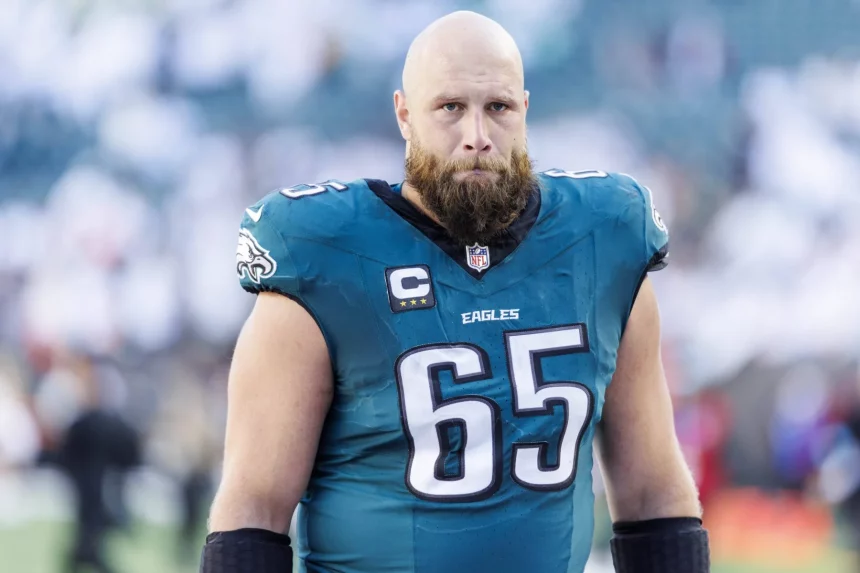 Lane Johnson in einem blaugrünen Trikot der Philadelphia Eagles mit der Nummer 65, vermutlich Lane Johnson, steht auf dem Spielfeld. Er hat einen dichten Bart und eine Glatze. Auf der Uniform des Spielers ist ein Kapitänsabzeichen zu sehen. Im Hintergrund sind eine verschwommene Menge und ein Teil des Stadions zu sehen, die möglicherweise seine Vertragsverlängerung feiern. Diese Beschreibung wurde mit der FootballR KI automatisch generiert.