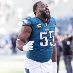 Ein Footballspieler, Brandon Graham, in einem blaugrünen Trikot mit der Nummer 55 und einem Kapitänsabzeichen, lächelt breit auf dem Spielfeld. Er gestikuliert mit seiner rechten Hand vor dem Hintergrund verschwommenen Stadionlichts, anderer Spieler und jubelnder Zuschauer. Diese Beschreibung wurde mit der FootballR KI automatisch generiert.