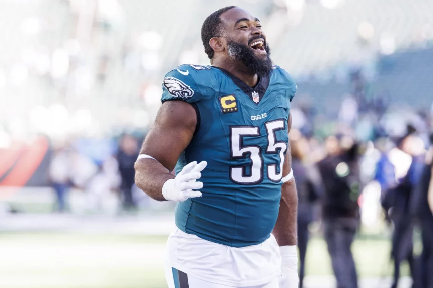 Ein Footballspieler, Brandon Graham, in einem blaugrünen Trikot mit der Nummer 55 und einem Kapitänsabzeichen, lächelt breit auf dem Spielfeld. Er gestikuliert mit seiner rechten Hand vor dem Hintergrund verschwommenen Stadionlichts, anderer Spieler und jubelnder Zuschauer. Diese Beschreibung wurde mit der FootballR KI automatisch generiert.