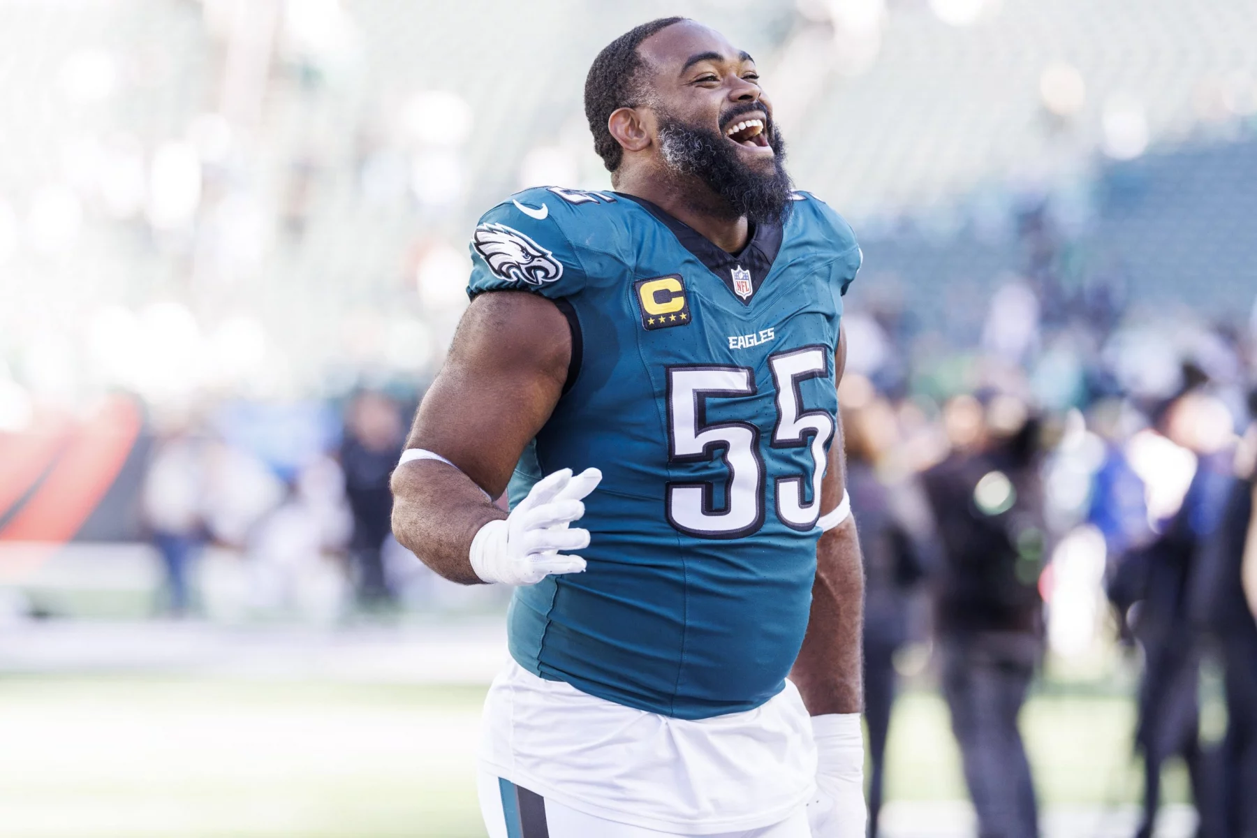 Ein Footballspieler, Brandon Graham, in einem blaugrünen Trikot mit der Nummer 55 und einem Kapitänsabzeichen, lächelt breit auf dem Spielfeld. Er gestikuliert mit seiner rechten Hand vor dem Hintergrund verschwommenen Stadionlichts, anderer Spieler und jubelnder Zuschauer. Diese Beschreibung wurde mit der FootballR KI automatisch generiert.