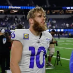 Cooper Kupp mit Bart, einem weißen Trikot mit der Nummer 10 und dem NFL-Logo steht auf einem Spielfeld. Im Hintergrund sind Fans und Stadionsitze zu sehen. Der Spieler wirkt konzentriert und leicht verschwitzt, mit zerzausten Haaren, nach einem scheinbaren Spiel. Diese Beschreibung wurde mit der FootballR KI automatisch generiert.