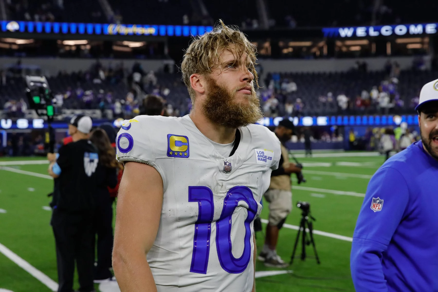 Cooper Kupp mit Bart, einem weißen Trikot mit der Nummer 10 und dem NFL-Logo steht auf einem Spielfeld. Im Hintergrund sind Fans und Stadionsitze zu sehen. Der Spieler wirkt konzentriert und leicht verschwitzt, mit zerzausten Haaren, nach einem scheinbaren Spiel. Diese Beschreibung wurde mit der FootballR KI automatisch generiert.
