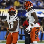 Ja'Marr Chase und Tee Higgins in orange-weißen Trikots stehen auf einem Spielfeld. Der linke Spieler, der Ja'Marr Chase ähnelt, lächelt unter seinem orange-schwarz gestreiften Helm. Rechts hält ein Spieler einen Football, der an Tee Higgins' Stil erinnert. Im Hintergrund ist eine verschwommene Menschenmenge zu sehen. Diese Beschreibung wurde mit der FootballR KI automatisch generiert.