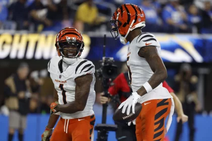 Ja'Marr Chase und Tee Higgins in orange-weißen Trikots stehen auf einem Spielfeld. Der linke Spieler, der Ja'Marr Chase ähnelt, lächelt unter seinem orange-schwarz gestreiften Helm. Rechts hält ein Spieler einen Football, der an Tee Higgins' Stil erinnert. Im Hintergrund ist eine verschwommene Menschenmenge zu sehen. Diese Beschreibung wurde mit der FootballR KI automatisch generiert.