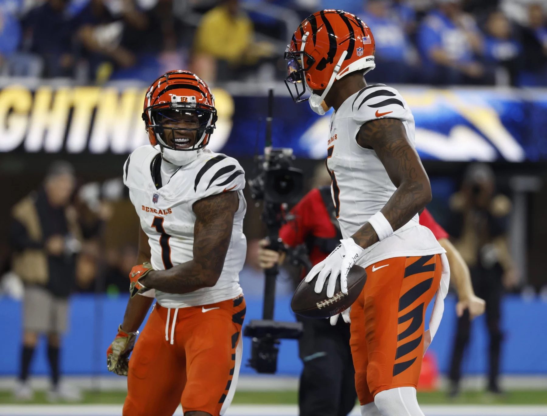 Ja'Marr Chase und Tee Higgins in orange-weißen Trikots stehen auf einem Spielfeld. Der linke Spieler, der Ja'Marr Chase ähnelt, lächelt unter seinem orange-schwarz gestreiften Helm. Rechts hält ein Spieler einen Football, der an Tee Higgins' Stil erinnert. Im Hintergrund ist eine verschwommene Menschenmenge zu sehen. Diese Beschreibung wurde mit der FootballR KI automatisch generiert.