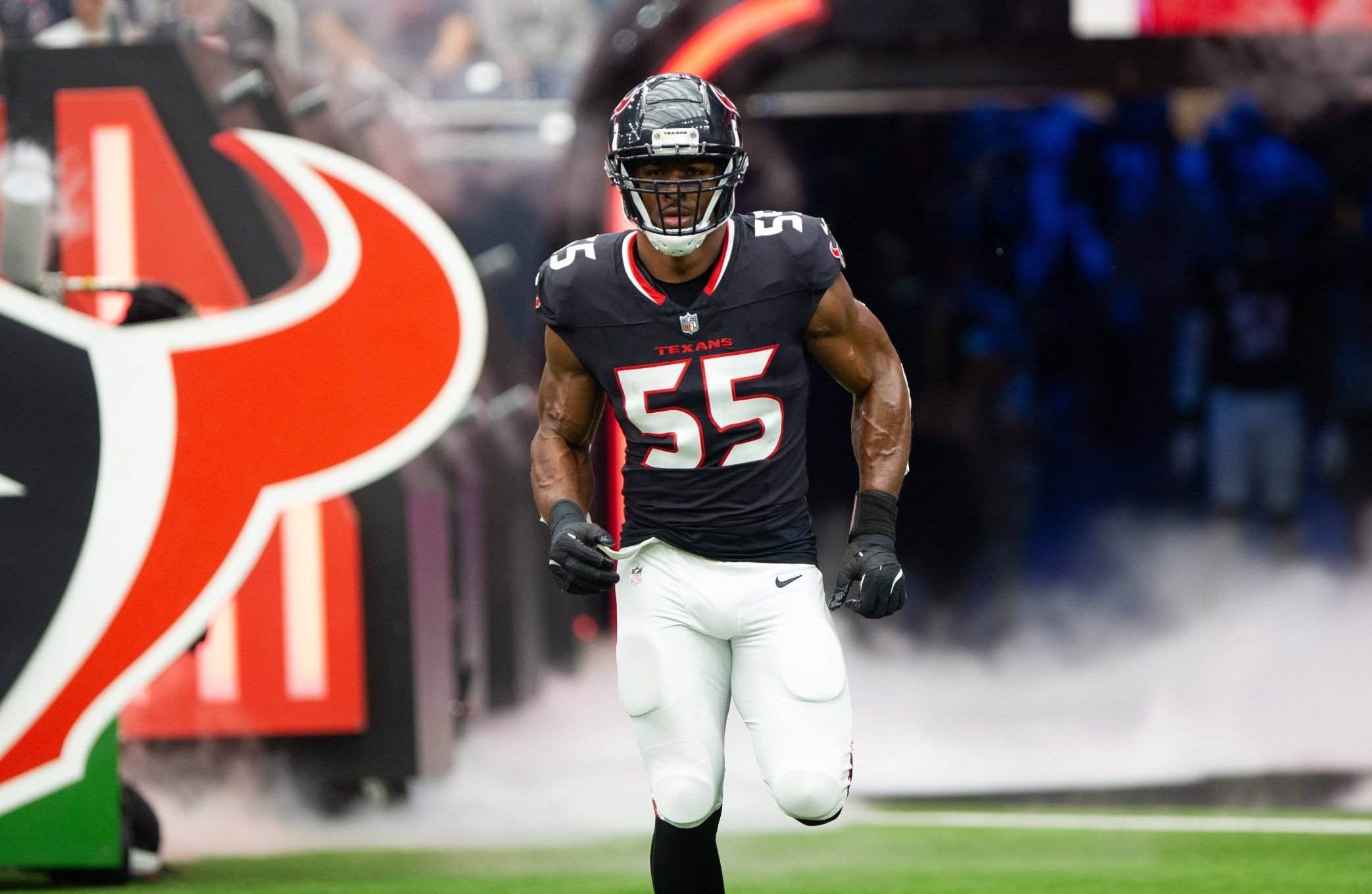 Danielle Hunter, in einem schwarzen Trikot mit der Nummer 55, stürmt aus einem Tunnel auf das Footballfeld. Hinter ihm quillt Rauch auf, während er Helm und weiße Hose anzieht. Ein großes Stierkopf-Logo kennzeichnet den Tunneleingang, im Hintergrund jubeln die Fans lautstark. Diese Beschreibung wurde mit der FootballR KI automatisch generiert.