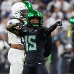 Ein Footballspieler, möglicherweise Tez Johnson, in schwarzer Uniform mit grünen Akzenten, zeigt mit beiden Händen. Sein glänzend grüner Helm glänzt im Stadionlicht. Ein weiterer Spieler in Weiß verweilt im Hintergrund, während die Fans die Tribünen füllen und die Spannung des Spiels auf dem Spielfeld einfangen. Diese Beschreibung wurde mit der FootballR KI automatisch generiert.