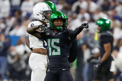 Ein Footballspieler, möglicherweise Tez Johnson, in schwarzer Uniform mit grünen Akzenten, zeigt mit beiden Händen. Sein glänzend grüner Helm glänzt im Stadionlicht. Ein weiterer Spieler in Weiß verweilt im Hintergrund, während die Fans die Tribünen füllen und die Spannung des Spiels auf dem Spielfeld einfangen. Diese Beschreibung wurde mit der FootballR KI automatisch generiert.