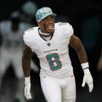 Jevon Holland in weißer Uniform der Miami Dolphins und blaukarierter Kappe, identifiziert als Jevon Holland, lächelt breit beim Laufen. Er trägt ein Trikot mit der Nummer 8. Im Hintergrund ist ein dunkler Tunnel zu sehen, in dessen Schatten ein anderer Spieler leicht zu erkennen ist. Diese Beschreibung wurde mit der FootballR KI automatisch generiert.